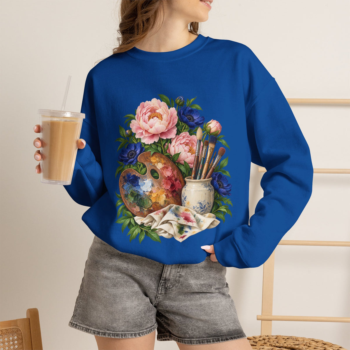 The Colors She Kept crewneck sweatshirt, a paint-covered artist palette beside a cream ceramic jar of brushes with pink peonies and deep blue flowers, image 28 of 28