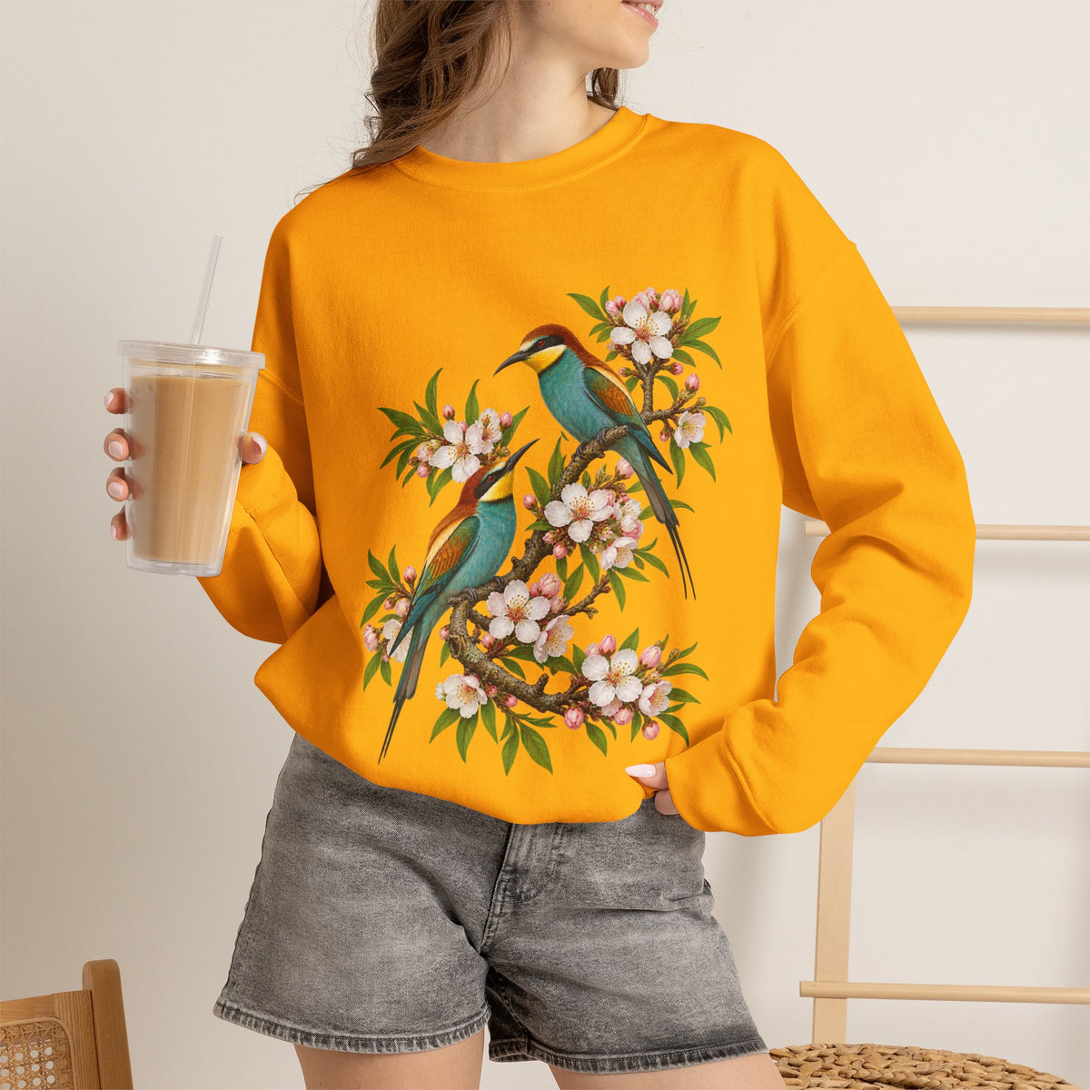 The Scenic Way Home sweatshirt — two birds with teal, golden-yellow and rust plumage perched among cream and blush blossoms, on a unisex crewneck