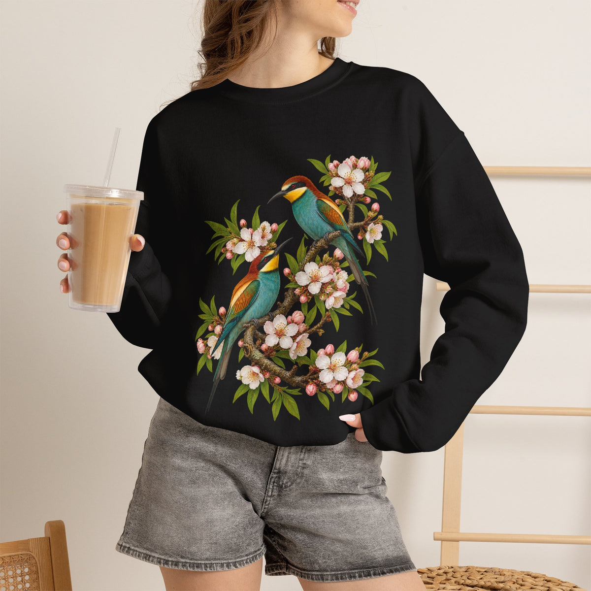 The Scenic Way Home sweatshirt — two birds with teal, golden-yellow and rust plumage perched among cream and blush blossoms, on a unisex crewneck