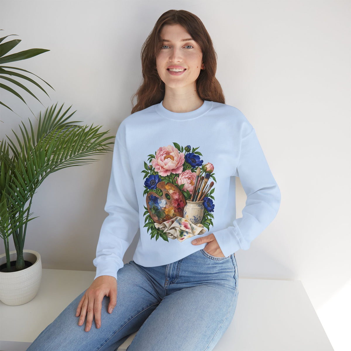 The Colors She Kept crewneck sweatshirt, a paint-covered artist palette beside a cream ceramic jar of brushes with pink peonies and deep blue flowers, image 17 of 28
