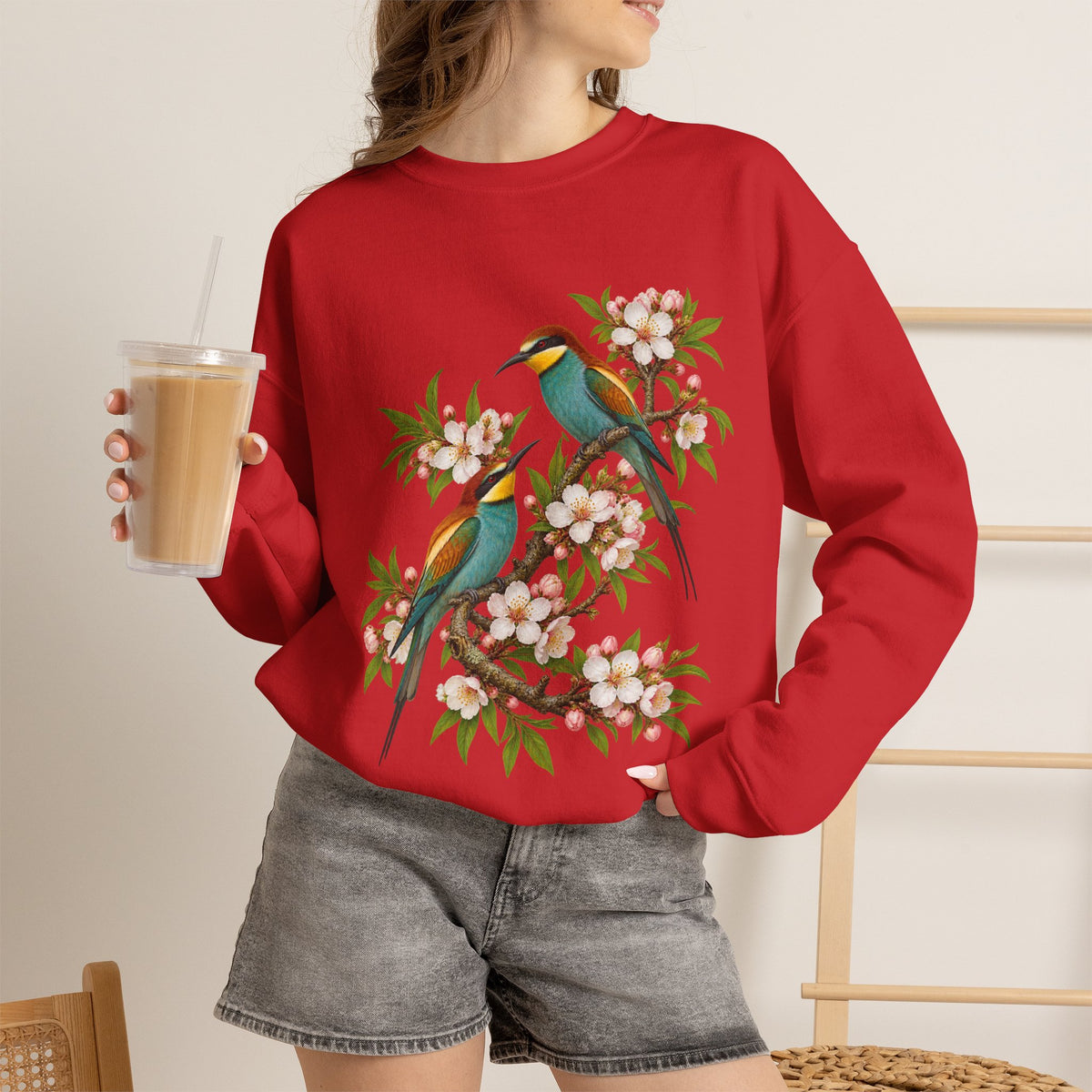 The Scenic Way Home sweatshirt — two birds with teal, golden-yellow and rust plumage perched among cream and blush blossoms, on a unisex crewneck