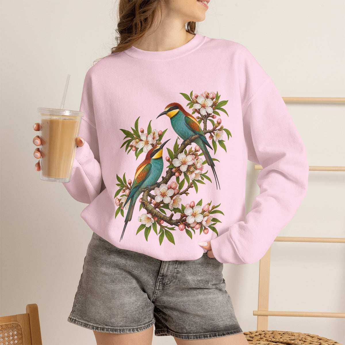 The Scenic Way Home sweatshirt — two birds with teal, golden-yellow and rust plumage perched among cream and blush blossoms, on a unisex crewneck