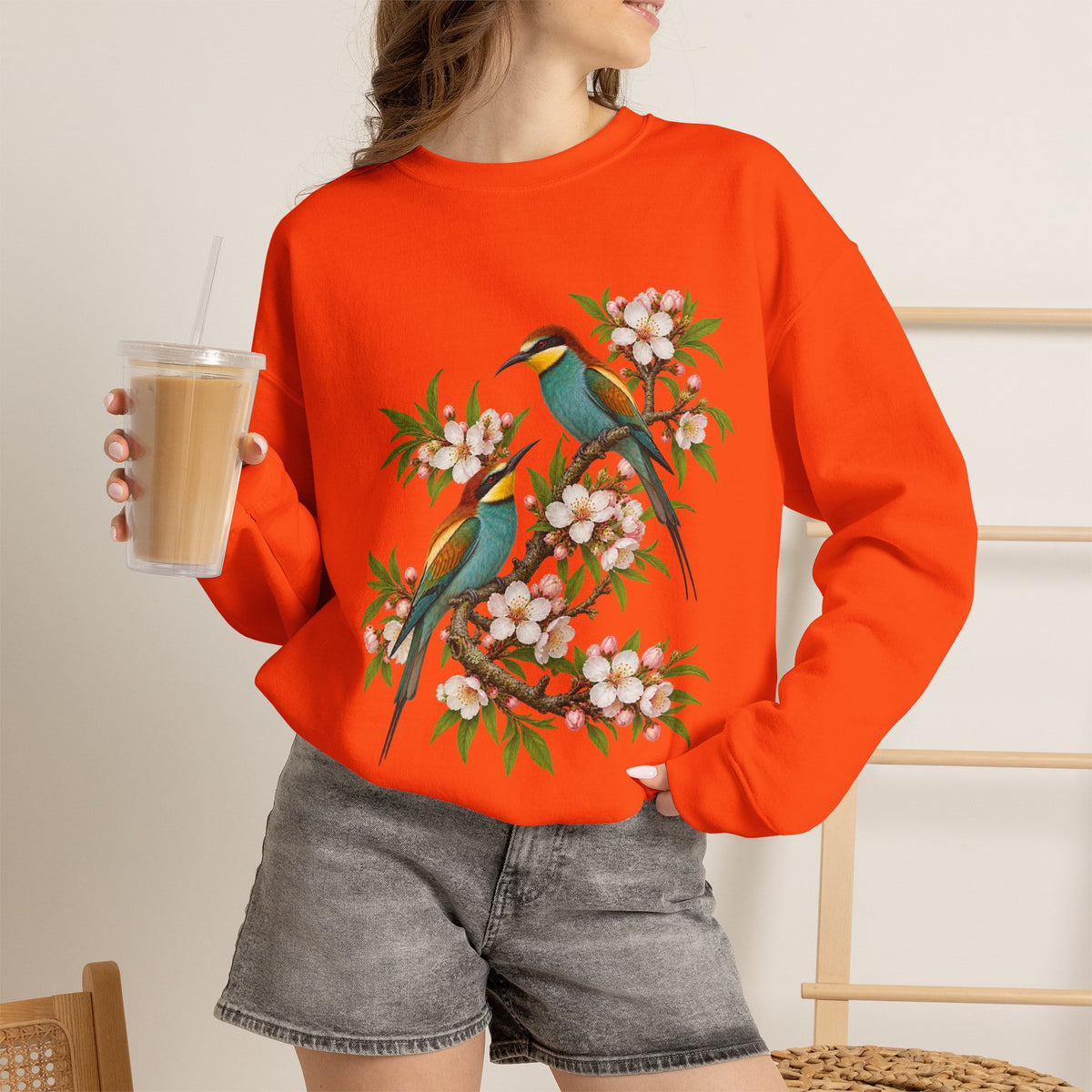 The Scenic Way Home sweatshirt — two birds with teal, golden-yellow and rust plumage perched among cream and blush blossoms, on a unisex crewneck