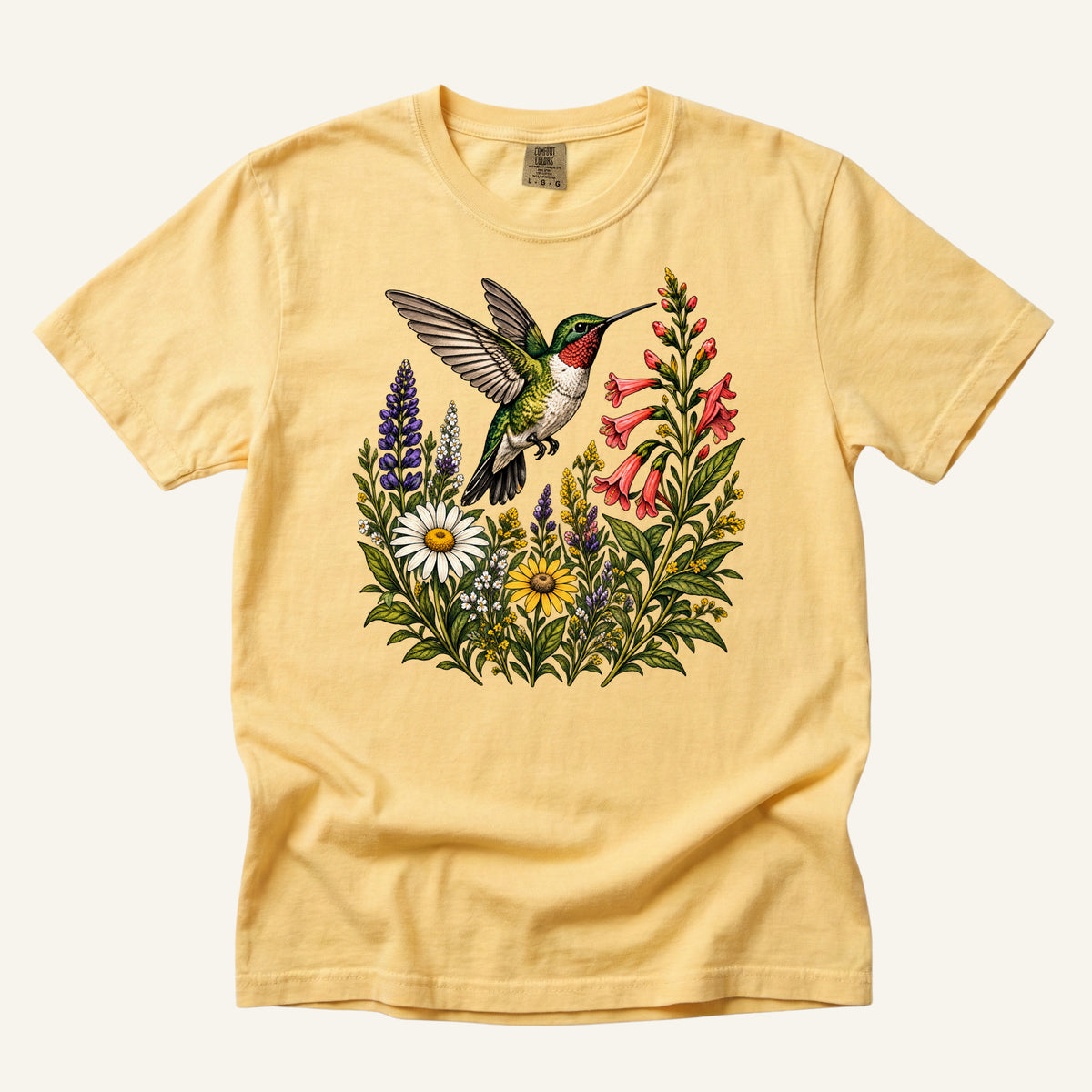 A Moment on Wings tee in Butter, a hummingbird hovering beside red trumpet blooms and purple wildflower spikes