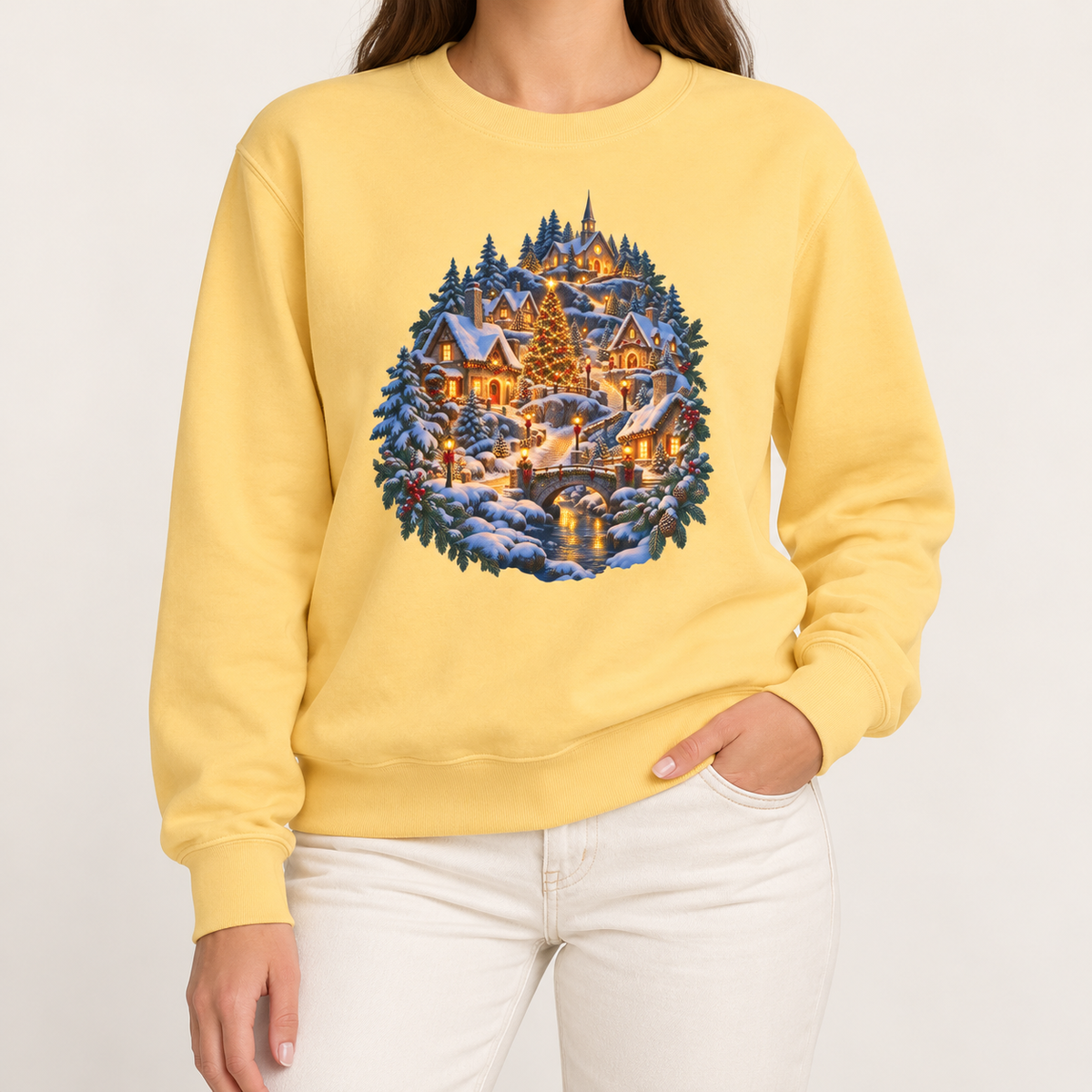 Small town Christmas sweatshirt in Butter: a snowy hillside village with a lit church, Christmas tree and stone bridge, on a soft pale yellow garment-dyed Comfort Colors crewneck