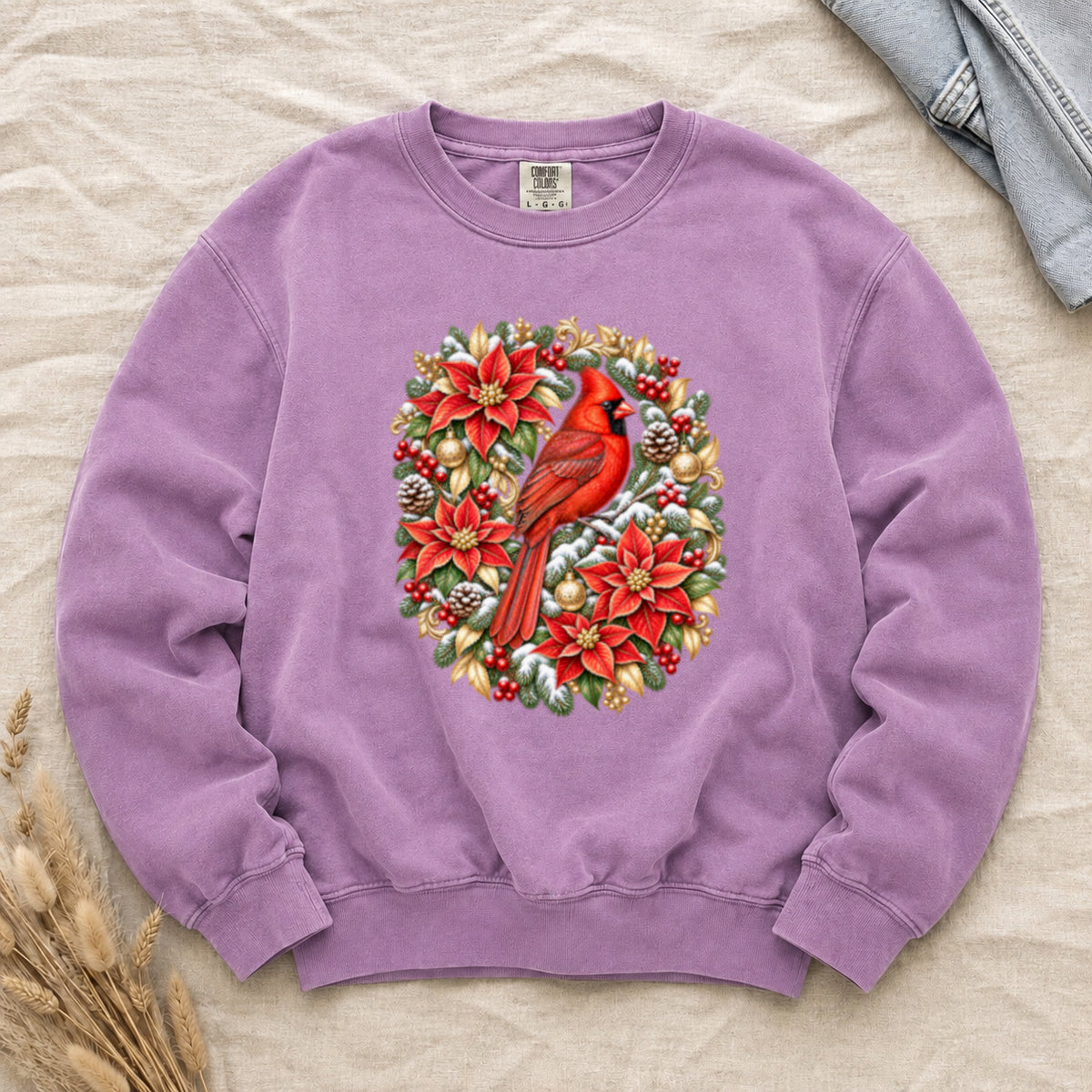 Northern Cardinal and Poinsettia Christmas Sweatshirt in Orchid — a Northern Cardinal perched at the centre of an oval wreath of red poinsettias, snow-dusted evergreen, gold leaves and ornaments, berries and pinecones
