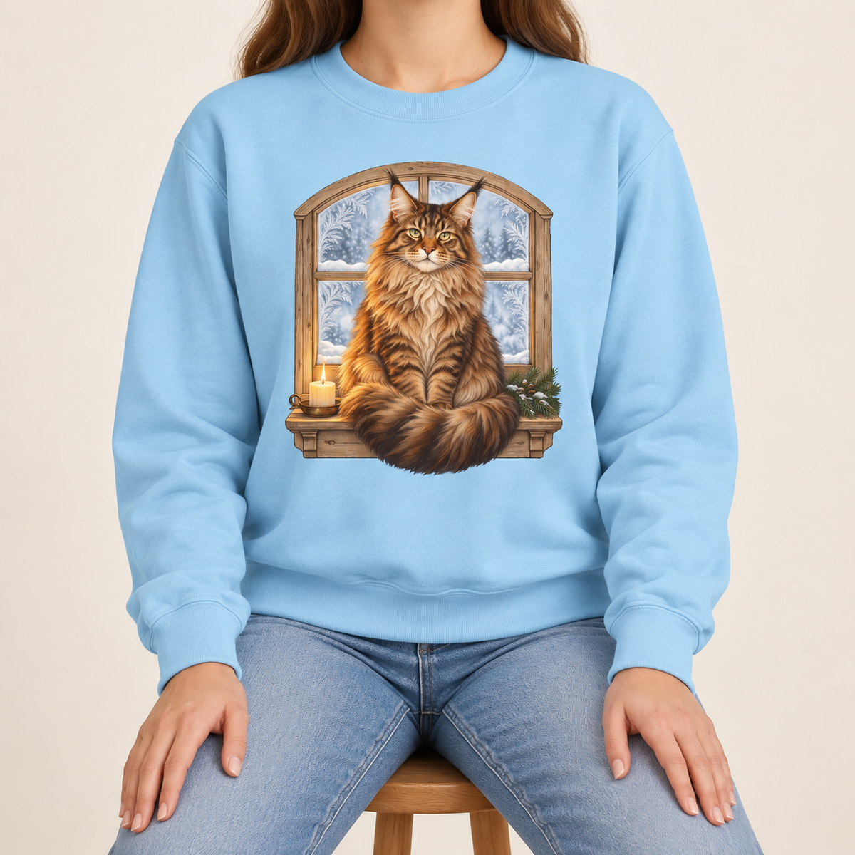Maine Coon at the window sweatshirt in Chambray – a tabby Maine Coon on a frosted windowsill with a candle and pine, snowy forest through an arched window, printed large on the chest of a Comfort Colors crewneck