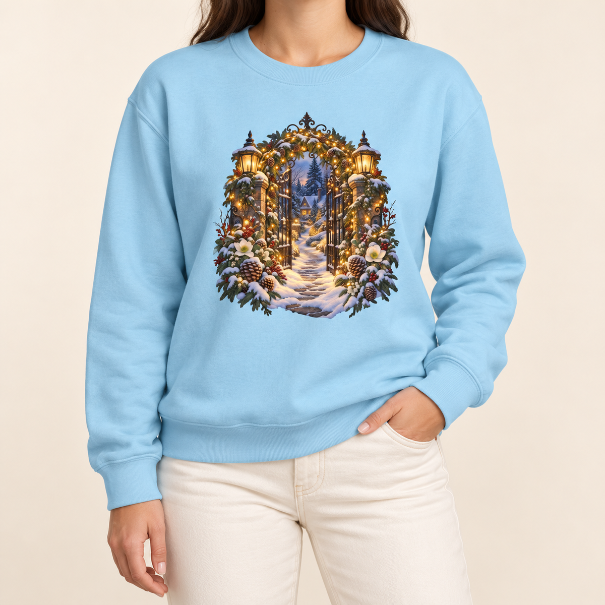 Christmas garden gate sweatshirt in Chambray: an open iron gate with lantern-topped pillars and a snowy path to a lit cottage, on a muted mid-blue garment-dyed Comfort Colors crewneck