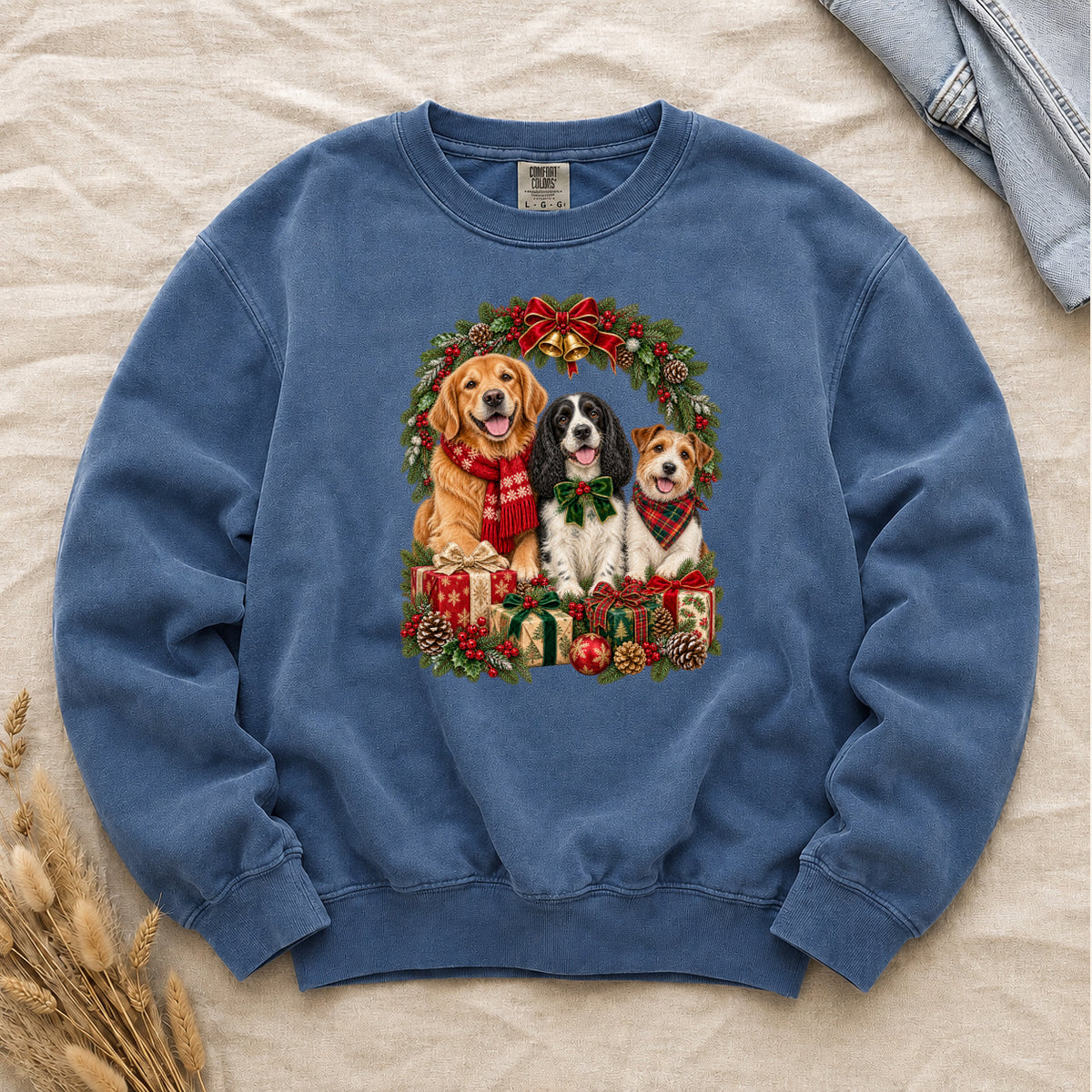 Three Dogs & Christmas Wreath Sweatshirt in Blue Jean — a golden retriever in a red snowflake scarf, a black-and-white spaniel in a green bow and a small tan-and-white terrier in a red plaid bandana, framed by an evergreen Christmas wreath with a tartan bow, a gold bell and wrapped gifts, printed large and centered on a washed light-mid blue garment-dyed Comfort Colors crewneck