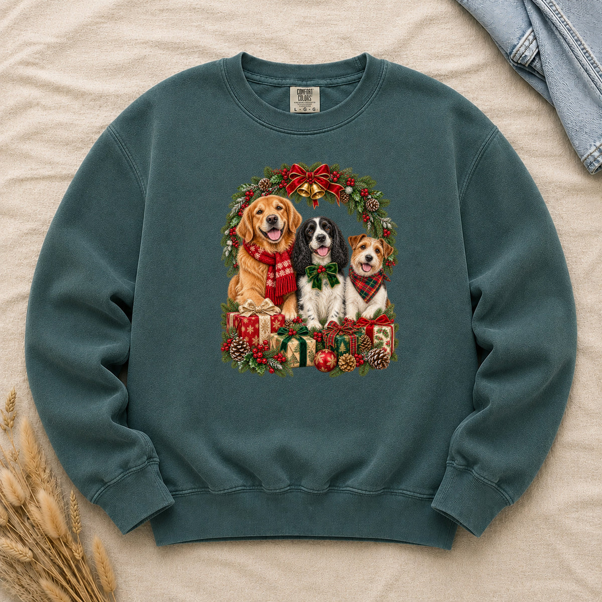 Three Dogs & Christmas Wreath Sweatshirt in Blue Spruce — a golden retriever in a red snowflake scarf, a black-and-white spaniel in a green bow and a small tan-and-white terrier in a red plaid bandana, framed by an evergreen Christmas wreath with a tartan bow, a gold bell and wrapped gifts, printed large and centered on a deep spruce green garment-dyed Comfort Colors crewneck