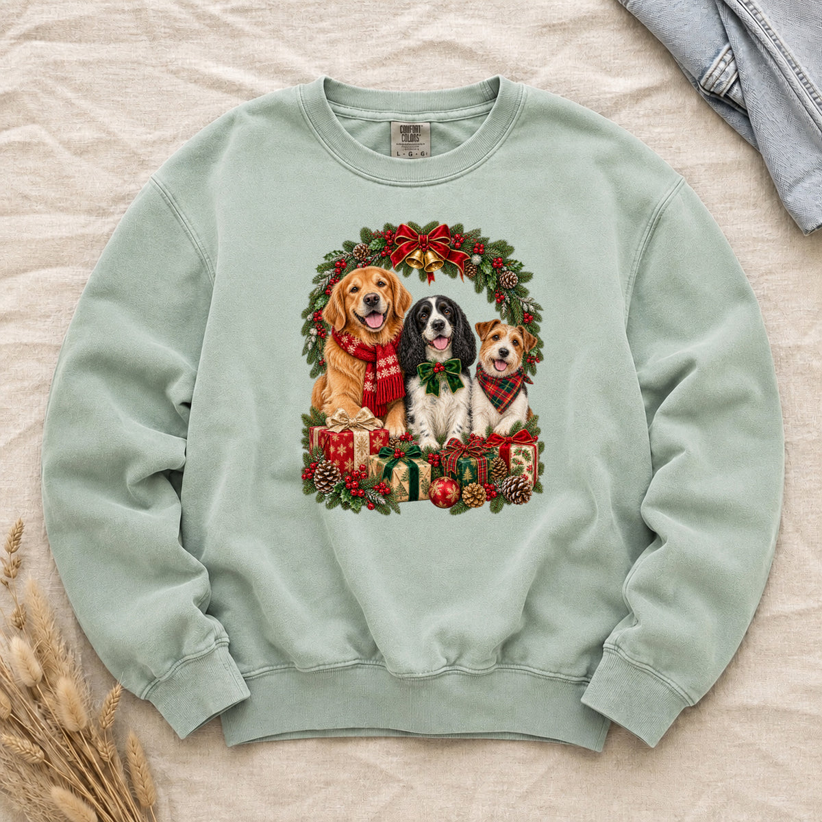 Three Dogs & Christmas Wreath Sweatshirt in Chalky Mint — a golden retriever in a red snowflake scarf, a black-and-white spaniel in a green bow and a small tan-and-white terrier in a red plaid bandana, framed by an evergreen Christmas wreath with a tartan bow, a gold bell and wrapped gifts, printed large and centered on a pale mint garment-dyed Comfort Colors crewneck