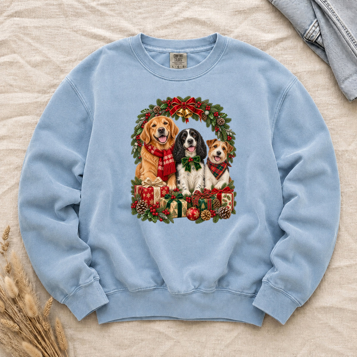 Three Dogs & Christmas Wreath Sweatshirt in Chambray — a golden retriever in a red snowflake scarf, a black-and-white spaniel in a green bow and a small tan-and-white terrier in a red plaid bandana, framed by an evergreen Christmas wreath with a tartan bow, a gold bell and wrapped gifts, printed large and centered on a pale chambray blue garment-dyed Comfort Colors crewneck
