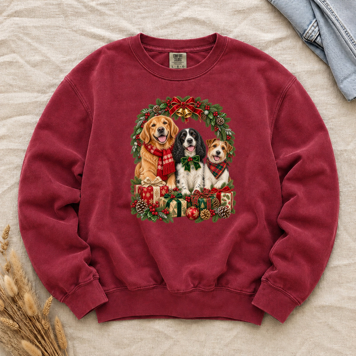 Three Dogs & Christmas Wreath Sweatshirt in Crimson — a golden retriever in a red snowflake scarf, a black-and-white spaniel in a green bow and a small tan-and-white terrier in a red plaid bandana, framed by an evergreen Christmas wreath with a tartan bow, a gold bell and wrapped gifts, printed large and centered on a deep crimson garment-dyed Comfort Colors crewneck