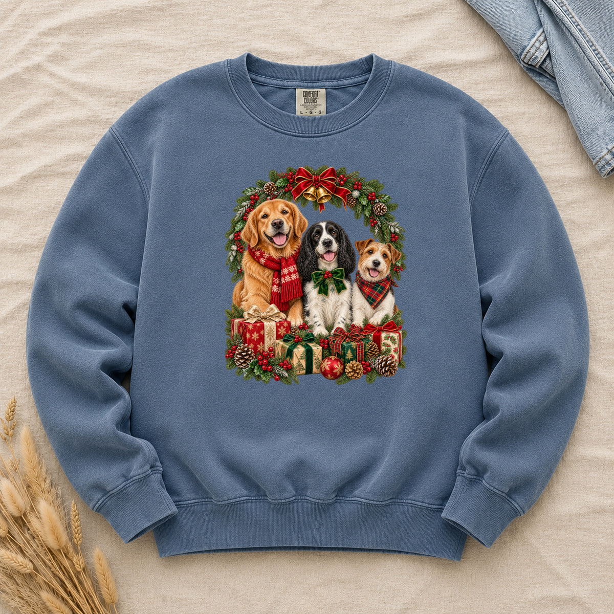 Three Dogs & Christmas Wreath Sweatshirt in Denim — a golden retriever in a red snowflake scarf, a black-and-white spaniel in a green bow and a small tan-and-white terrier in a red plaid bandana, framed by an evergreen Christmas wreath with a tartan bow, a gold bell and wrapped gifts, printed large and centered on a washed mid-blue garment-dyed Comfort Colors crewneck
