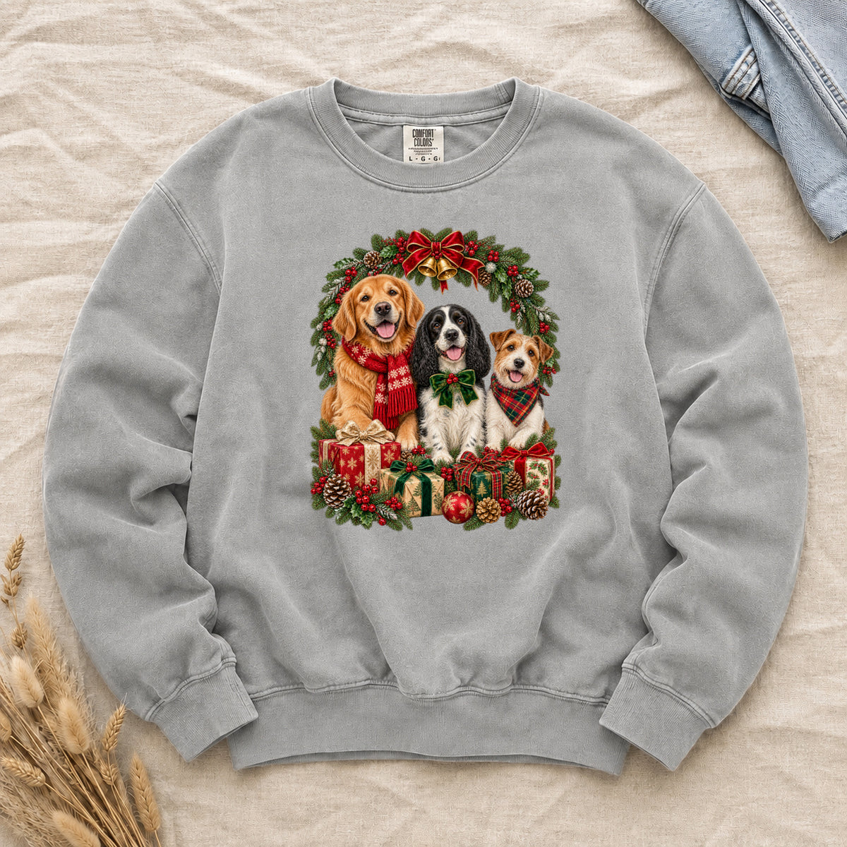Three Dogs & Christmas Wreath Sweatshirt in Grey — a golden retriever in a red snowflake scarf, a black-and-white spaniel in a green bow and a small tan-and-white terrier in a red plaid bandana, framed by an evergreen Christmas wreath with a tartan bow, a gold bell and wrapped gifts, printed large and centered on a soft grey garment-dyed Comfort Colors crewneck