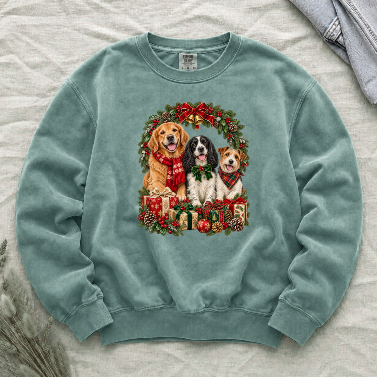 Three Dogs & Christmas Wreath Sweatshirt in Light Green — a golden retriever in a red snowflake scarf, a black-and-white spaniel in a green bow and a small tan-and-white terrier in a red plaid bandana, framed by an evergreen Christmas wreath with a tartan bow, a gold bell and wrapped gifts, printed large and centered on a pale green garment-dyed Comfort Colors crewneck