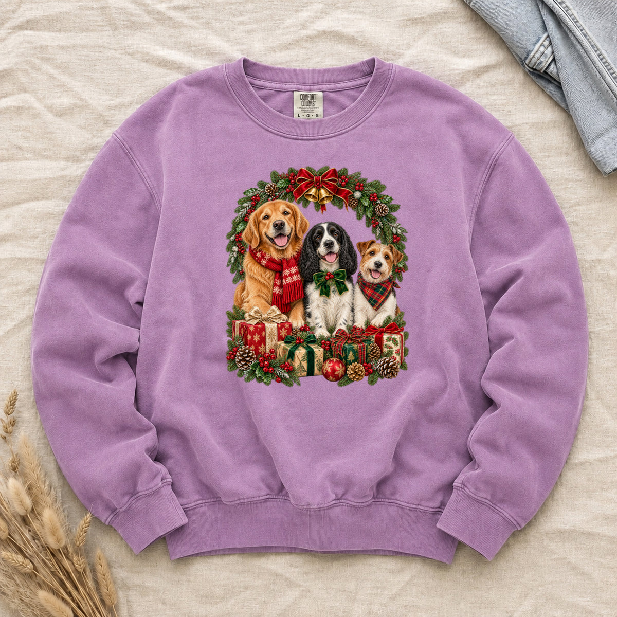 Three Dogs & Christmas Wreath Sweatshirt in Orchid — a golden retriever in a red snowflake scarf, a black-and-white spaniel in a green bow and a small tan-and-white terrier in a red plaid bandana, framed by an evergreen Christmas wreath with a tartan bow, a gold bell and wrapped gifts, printed large and centered on a soft lilac garment-dyed Comfort Colors crewneck