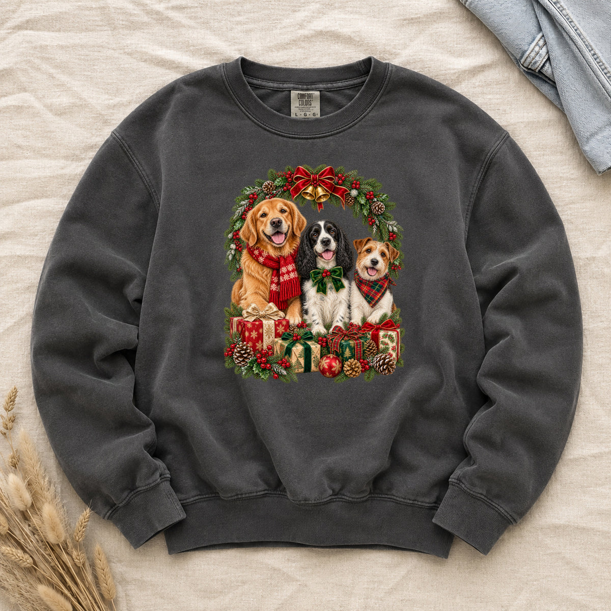 Three Dogs & Christmas Wreath Sweatshirt in Pepper — a golden retriever in a red snowflake scarf, a black-and-white spaniel in a green bow and a small tan-and-white terrier in a red plaid bandana, framed by an evergreen Christmas wreath with a tartan bow, a gold bell and wrapped gifts, printed large and centered on a deep charcoal garment-dyed Comfort Colors crewneck