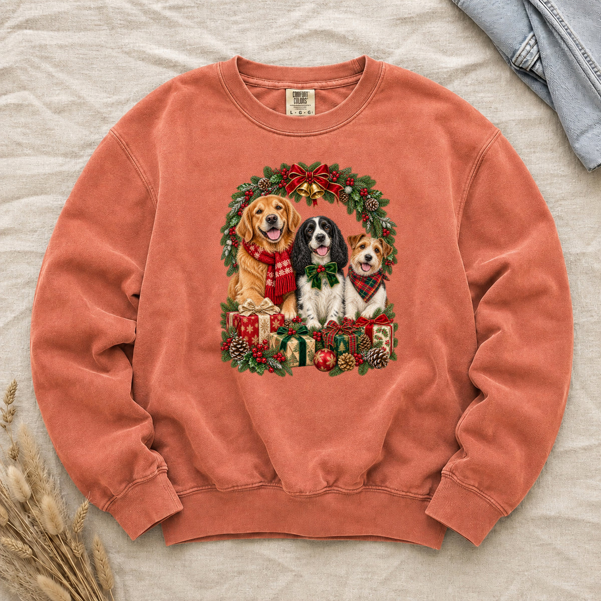 Three Dogs & Christmas Wreath Sweatshirt in Terracotta — a golden retriever in a red snowflake scarf, a black-and-white spaniel in a green bow and a small tan-and-white terrier in a red plaid bandana, framed by an evergreen Christmas wreath with a tartan bow, a gold bell and wrapped gifts, printed large and centered on a warm terracotta garment-dyed Comfort Colors crewneck