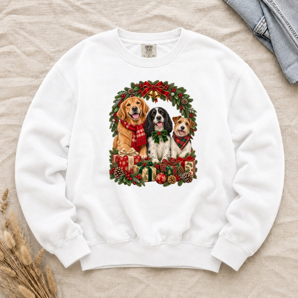 Three Dogs & Christmas Wreath Sweatshirt in White — a golden retriever in a red snowflake scarf, a black-and-white spaniel in a green bow and a small tan-and-white terrier in a red plaid bandana, framed by an evergreen Christmas wreath with a tartan bow, a gold bell and wrapped gifts, printed large and centered on a white garment-dyed Comfort Colors crewneck