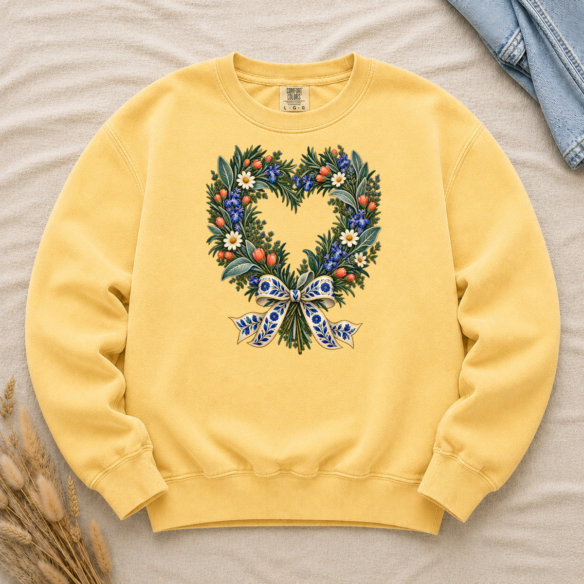 The Door with the Blue Bow floral heart wreath sweatshirt in Butter, leafy branches curving into a heart with cream daisies, blue flowers and red-orange berries, gathered at the base by a blue-and-gold patterned bow