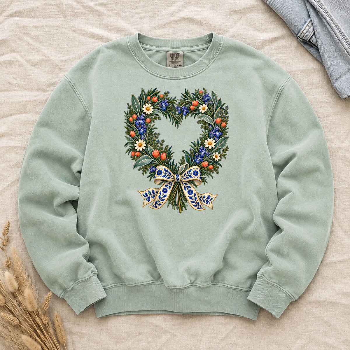 The Door with the Blue Bow floral heart wreath sweatshirt in Chalky Mint, leafy branches curving into a heart with cream daisies, blue flowers and red-orange berries, gathered at the base by a blue-and-gold patterned bow