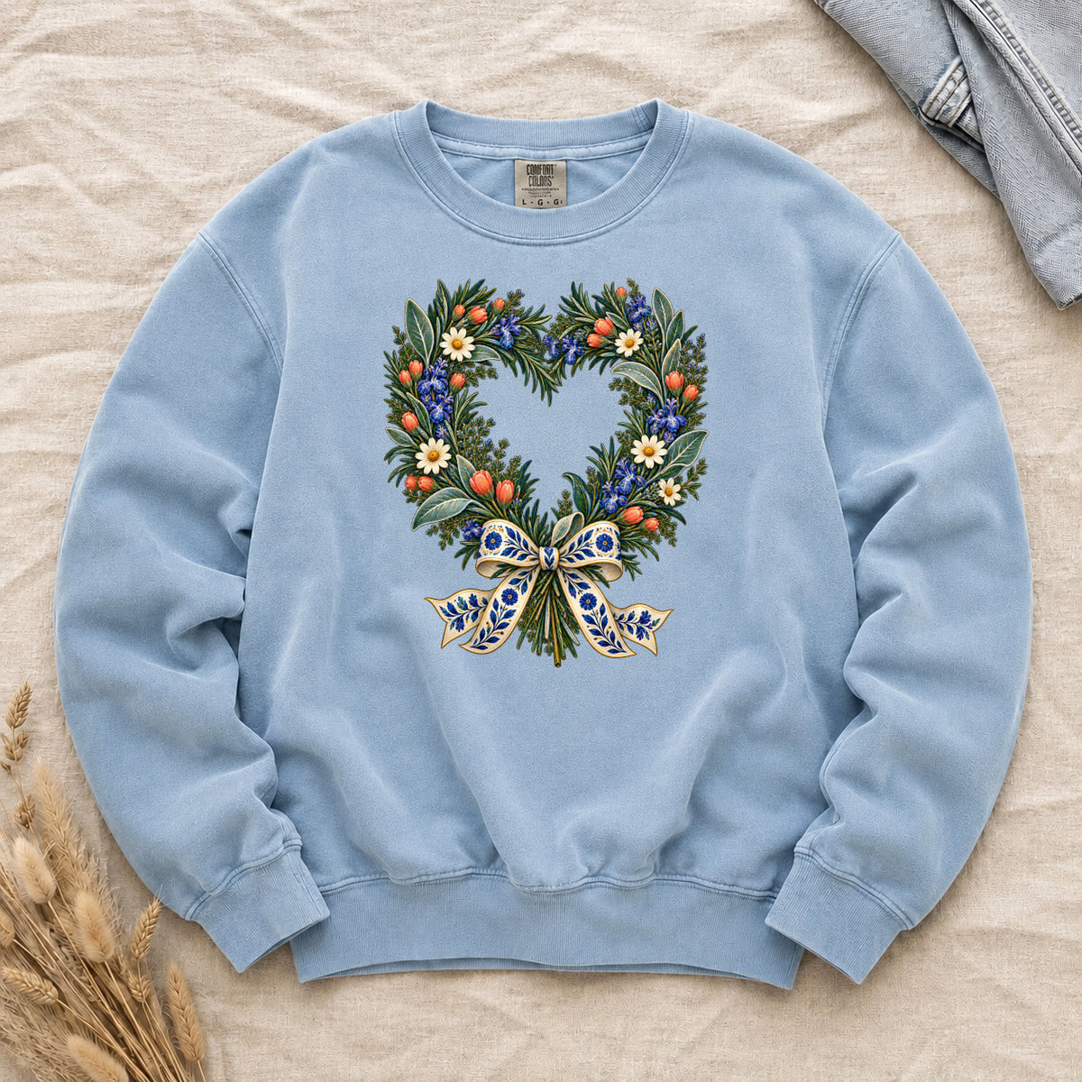 The Door with the Blue Bow floral heart wreath sweatshirt in Chambray, leafy branches curving into a heart with cream daisies, blue flowers and red-orange berries, gathered at the base by a blue-and-gold patterned bow