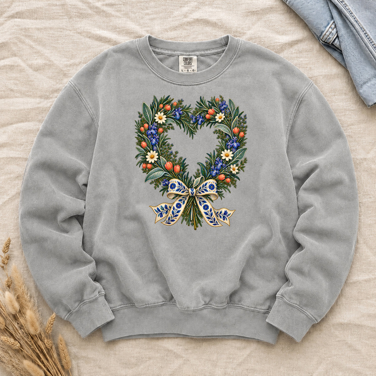 The Door with the Blue Bow floral heart wreath sweatshirt in Grey, leafy branches curving into a heart with cream daisies, blue flowers and red-orange berries, gathered at the base by a blue-and-gold patterned bow