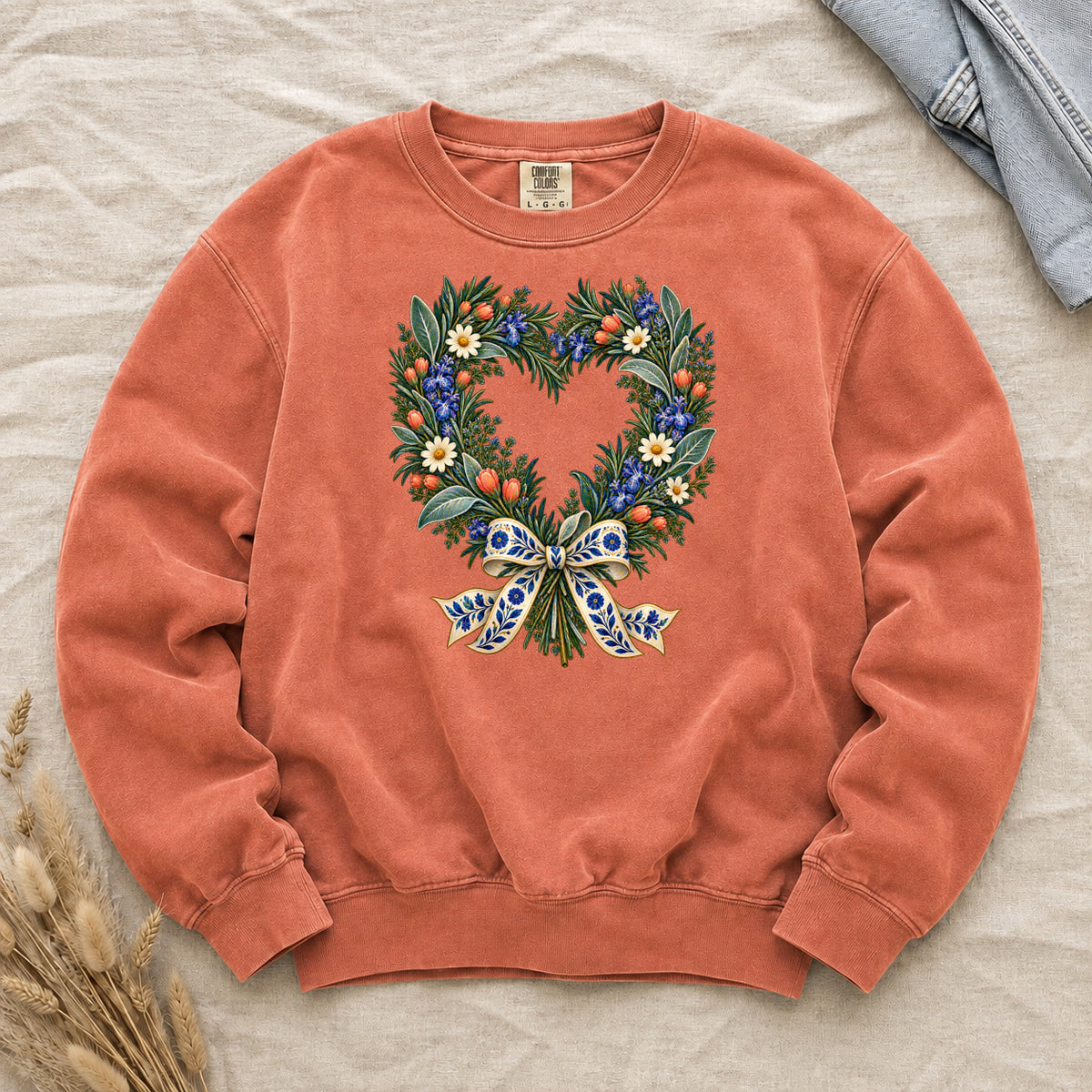 The Door with the Blue Bow floral heart wreath sweatshirt in Terracotta, leafy branches curving into a heart with cream daisies, blue flowers and red-orange berries, gathered at the base by a blue-and-gold patterned bow