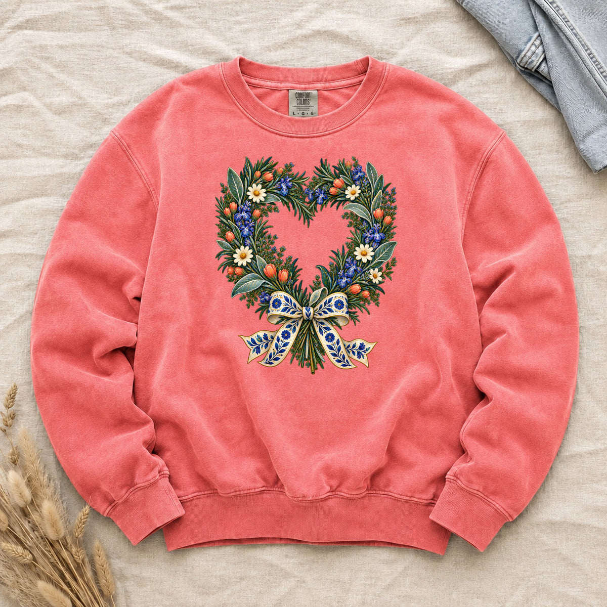 The Door with the Blue Bow floral heart wreath sweatshirt in Watermelon, leafy branches curving into a heart with cream daisies, blue flowers and red-orange berries, gathered at the base by a blue-and-gold patterned bow
