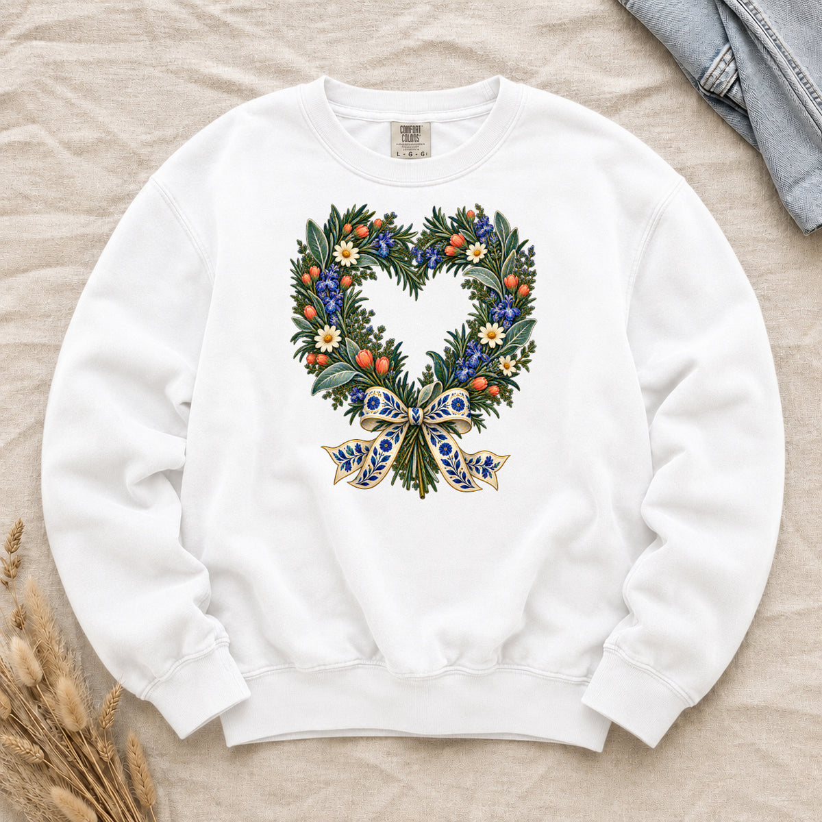 The Door with the Blue Bow floral heart wreath sweatshirt in White, leafy branches curving into a heart with cream daisies, blue flowers and red-orange berries, gathered at the base by a blue-and-gold patterned bow
