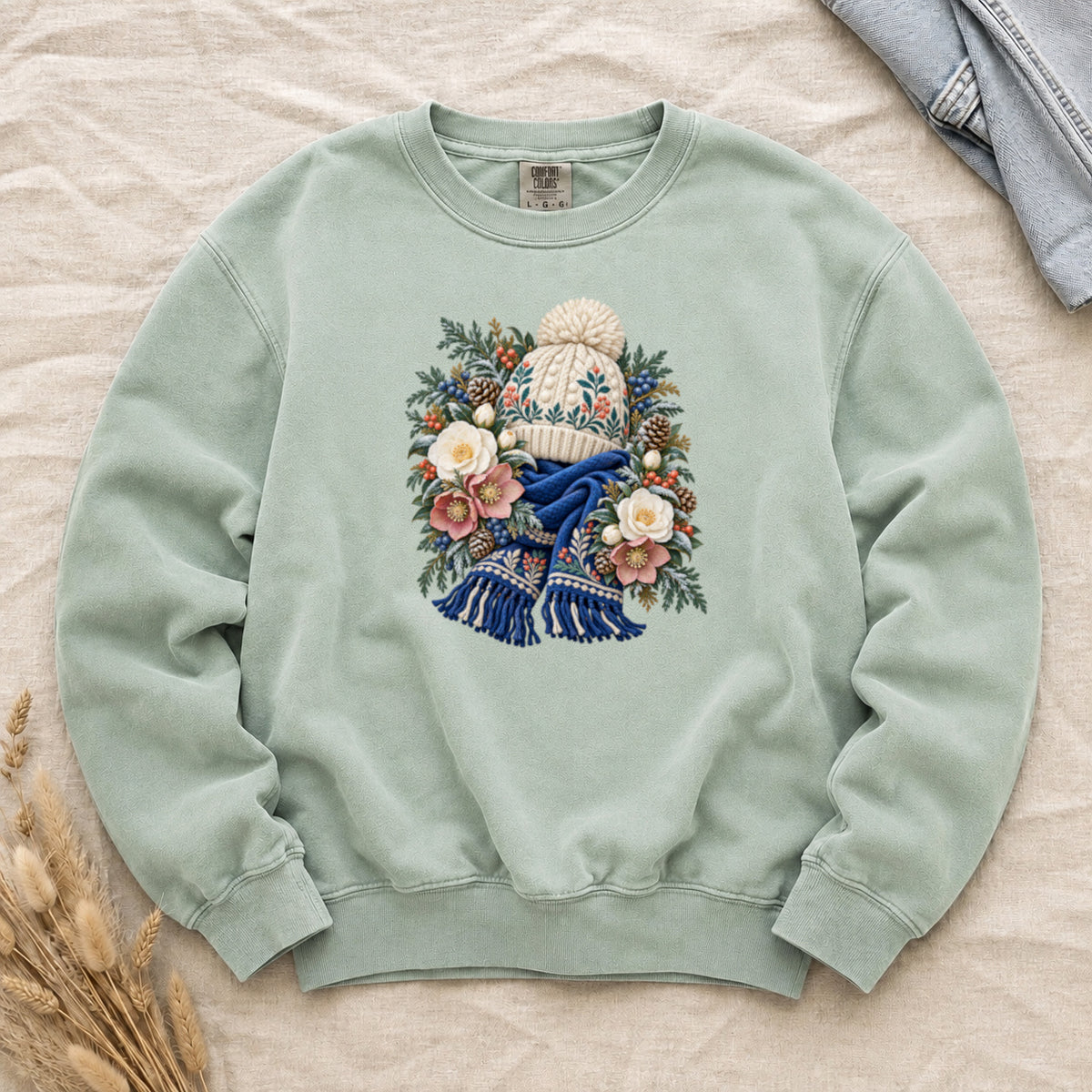 Floral Knit Hat & Scarf Sweatshirt in Chalky Mint — a cream pom-pom hat patterned in teal and coral above a deep blue fringed scarf, framed by evergreen, pinecones, red berries and cream roses