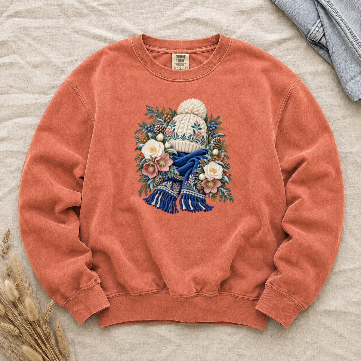 Floral Knit Hat & Scarf Sweatshirt in Terracotta — a cream pom-pom hat patterned in teal and coral above a deep blue fringed scarf, framed by evergreen, pinecones, red berries and cream roses