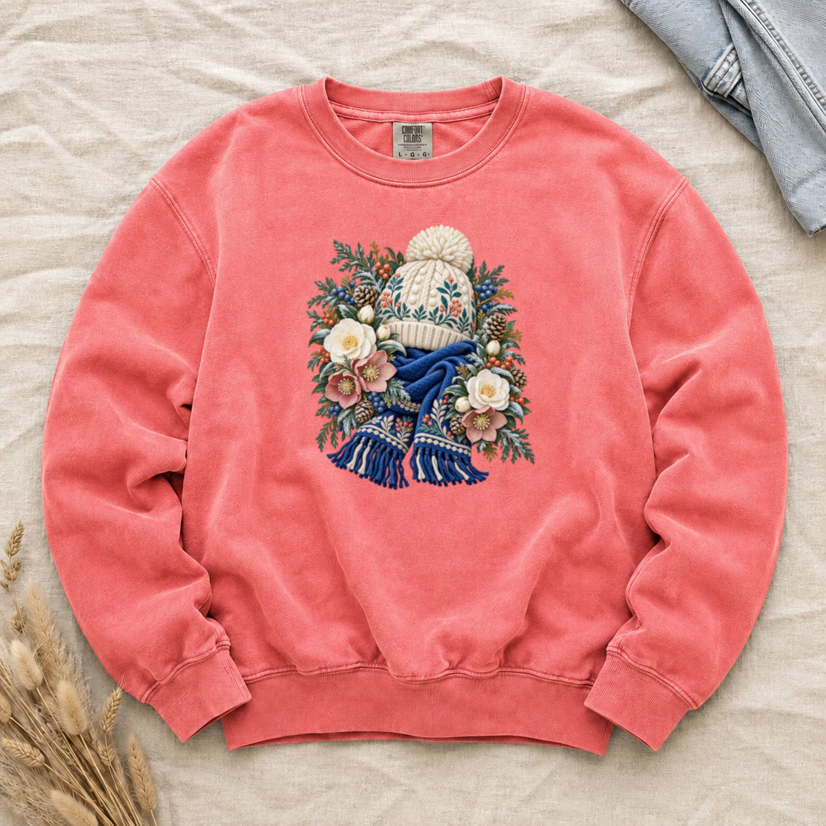 Floral Knit Hat & Scarf Sweatshirt in Watermelon — a cream pom-pom hat patterned in teal and coral above a deep blue fringed scarf, framed by evergreen, pinecones, red berries and cream roses