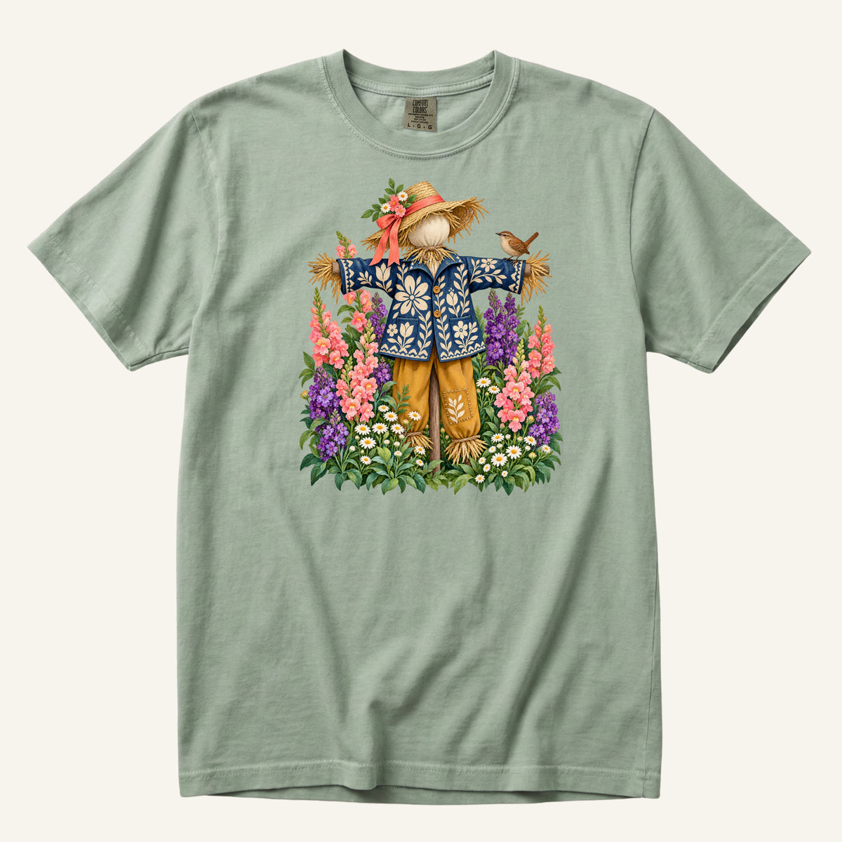 He Let Them Stay T-Shirt in Bay — a friendly scarecrow in a floral blue jacket with a small bird on his arm among garden flowers, sizes S to 4XL