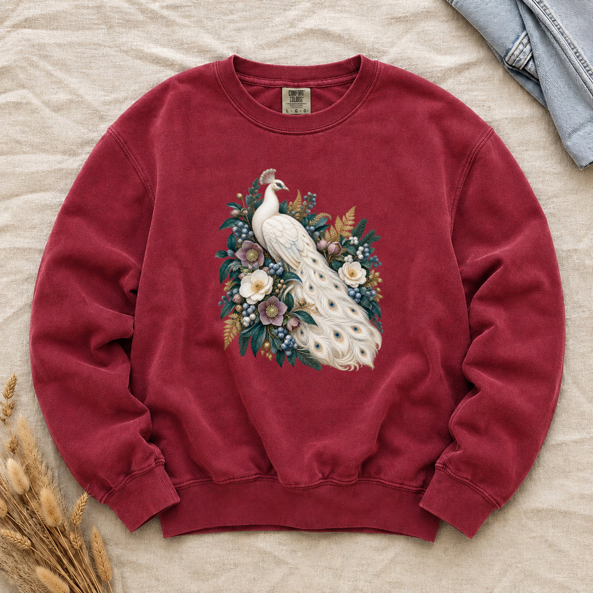 Ivory Peacock and Floral Sweatshirt in Crimson — an ivory peacock among cream and dusty purple flowers, blue berries and deep green leaves, its long tail curving down through the arrangement with small blue and gold markings on the pale feathers