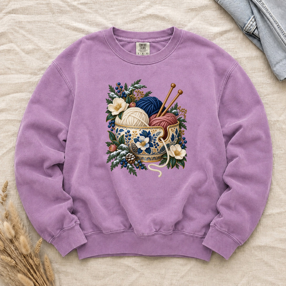 Knitting Yarn & Winter Botanicals Sweatshirt in Orchid — cream, deep blue and dusty rose yarn in an ornate blue-and-gold bowl with wooden knitting needles above it, among evergreen, pinecones, blue berries and cream flowers