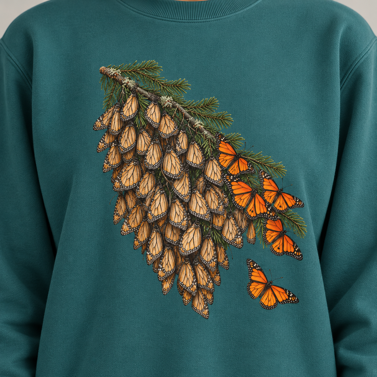 Close-up of the overwintering monarchs print on a Blue Spruce Comfort Colors crewneck - closed tan wings packed along a fir bough, open orange wings at the edge