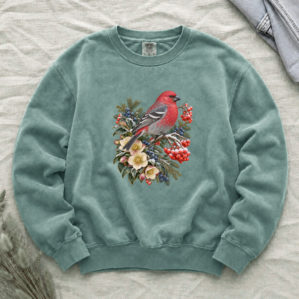 Red Bird & Winter Botanicals Sweatshirt in Light Green — a bright red bird with softly shaded gray wings and a long tail, perched on a branch among evergreen sprigs, red and blue berries and cream flowers