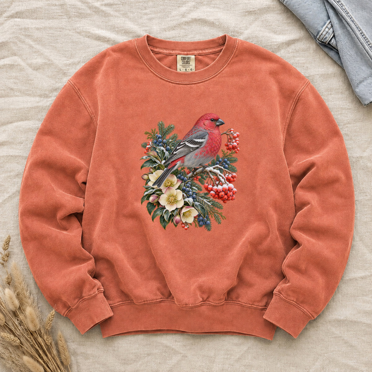 Red Bird & Winter Botanicals Sweatshirt in Terracotta — a bright red bird with softly shaded gray wings and a long tail, perched on a branch among evergreen sprigs, red and blue berries and cream flowers