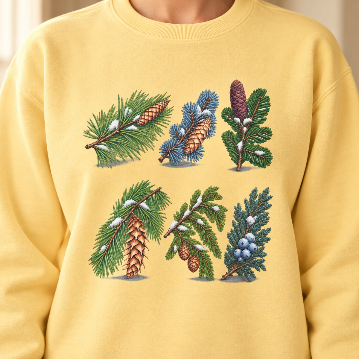 Close-up of the evergreen study print on a Butter Comfort Colors crewneck - pine, spruce, fir, Douglas fir, hemlock and juniper sprigs with cones and snow