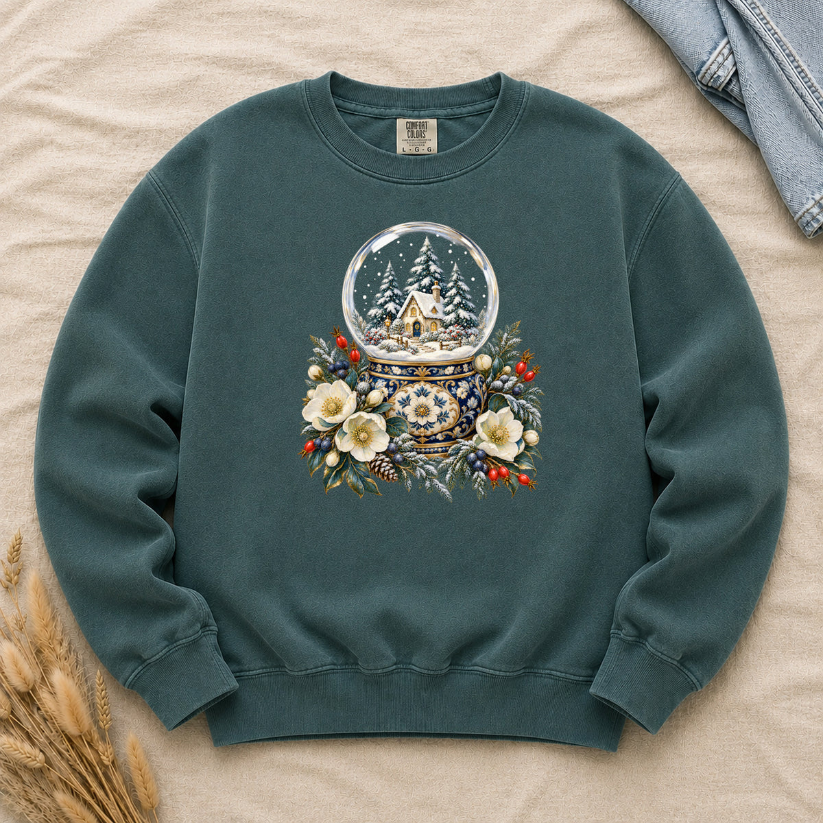 Snow Globe & Winter Florals Sweatshirt in Blue Spruce — a glass snow globe holding a snowy cottage and evergreens on an ornate navy-and-gold base, framed by cream blossoms, pinecones and red berries