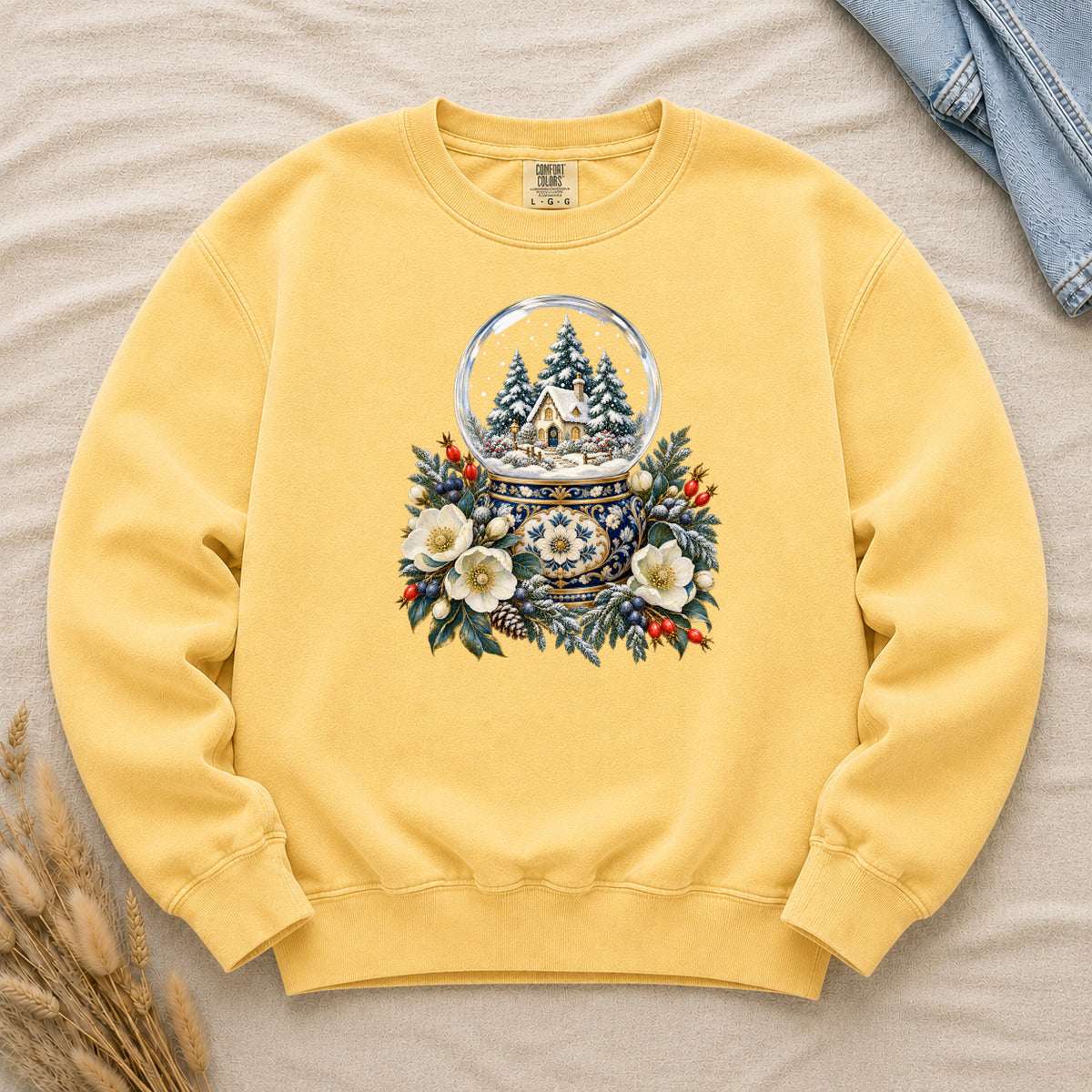Snow Globe & Winter Florals Sweatshirt in Butter — a glass snow globe holding a snowy cottage and evergreens on an ornate navy-and-gold base, framed by cream blossoms, pinecones and red berries