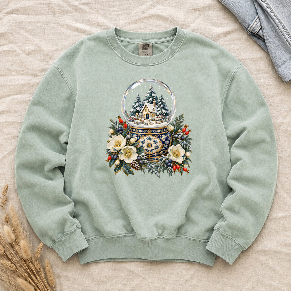 Snow Globe & Winter Florals Sweatshirt in Chalky Mint — a glass snow globe holding a snowy cottage and evergreens on an ornate navy-and-gold base, framed by cream blossoms, pinecones and red berries