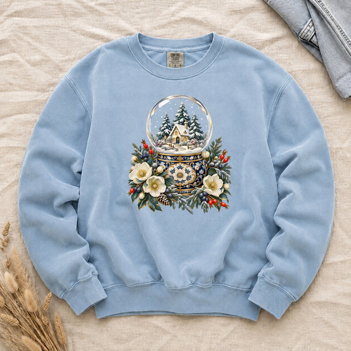 Snow Globe & Winter Florals Sweatshirt in Chambray — a glass snow globe holding a snowy cottage and evergreens on an ornate navy-and-gold base, framed by cream blossoms, pinecones and red berries