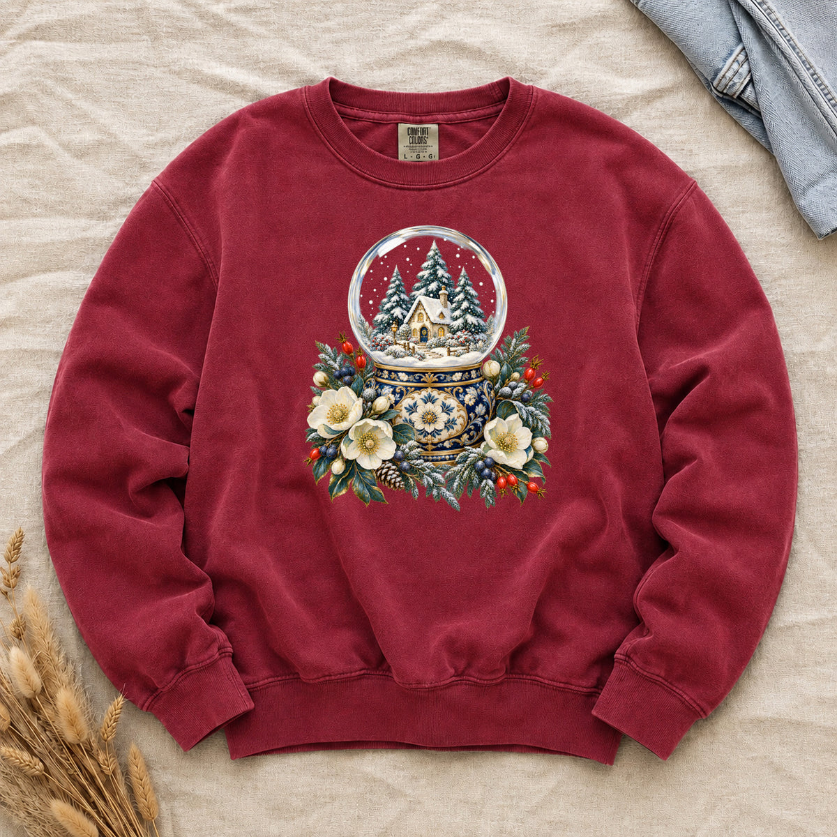 Snow Globe & Winter Florals Sweatshirt in Crimson — a glass snow globe holding a snowy cottage and evergreens on an ornate navy-and-gold base, framed by cream blossoms, pinecones and red berries