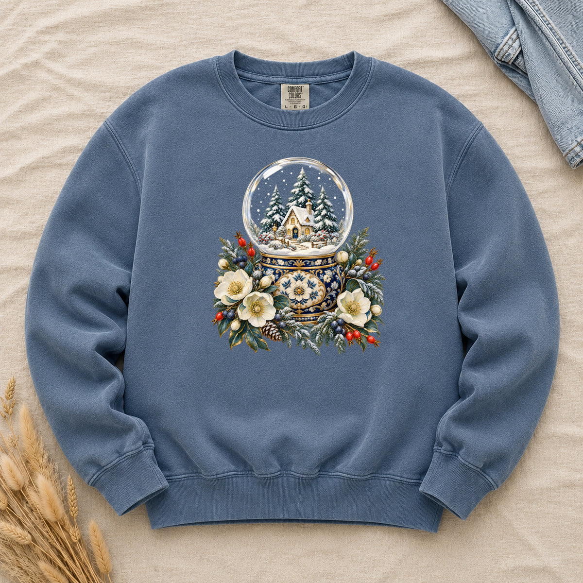 Snow Globe & Winter Florals Sweatshirt in Denim — a glass snow globe holding a snowy cottage and evergreens on an ornate navy-and-gold base, framed by cream blossoms, pinecones and red berries