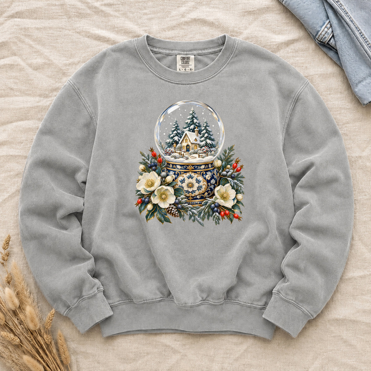 Snow Globe & Winter Florals Sweatshirt in Grey — a glass snow globe holding a snowy cottage and evergreens on an ornate navy-and-gold base, framed by cream blossoms, pinecones and red berries