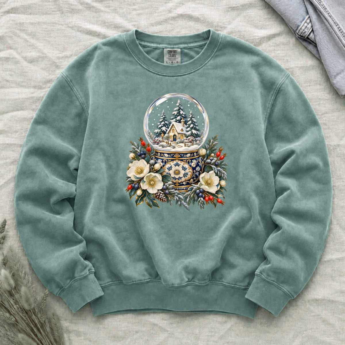 Snow Globe & Winter Florals Sweatshirt in Light Green — a glass snow globe holding a snowy cottage and evergreens on an ornate navy-and-gold base, framed by cream blossoms, pinecones and red berries
