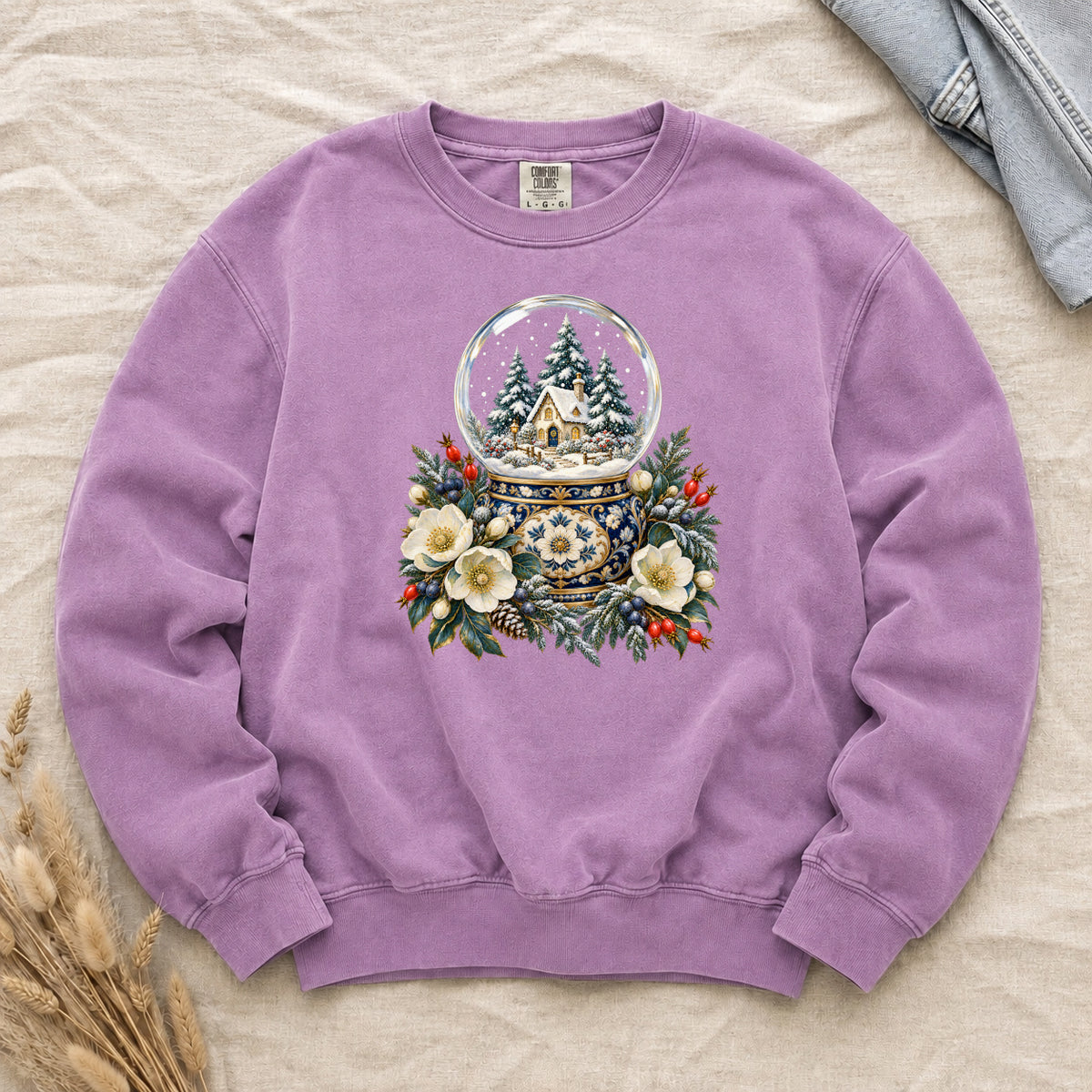 Snow Globe & Winter Florals Sweatshirt in Orchid — a glass snow globe holding a snowy cottage and evergreens on an ornate navy-and-gold base, framed by cream blossoms, pinecones and red berries